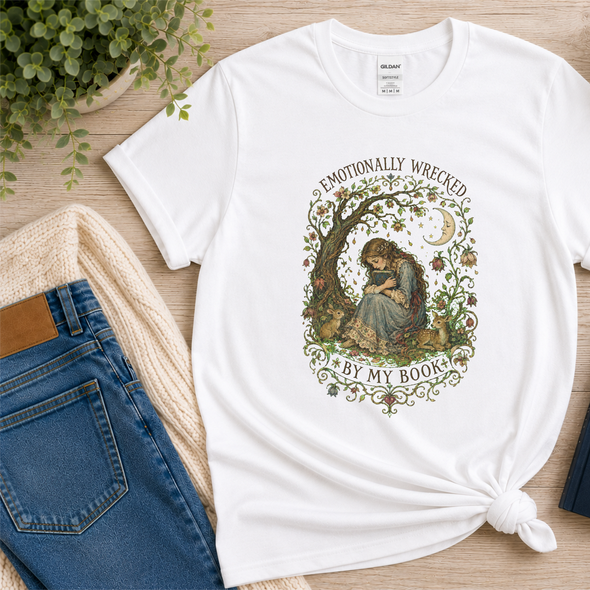 White T-shirt laid flat with a reader beneath a tree, flowers and a crescent moon and the words “Emotionally Wrecked by My Book.”