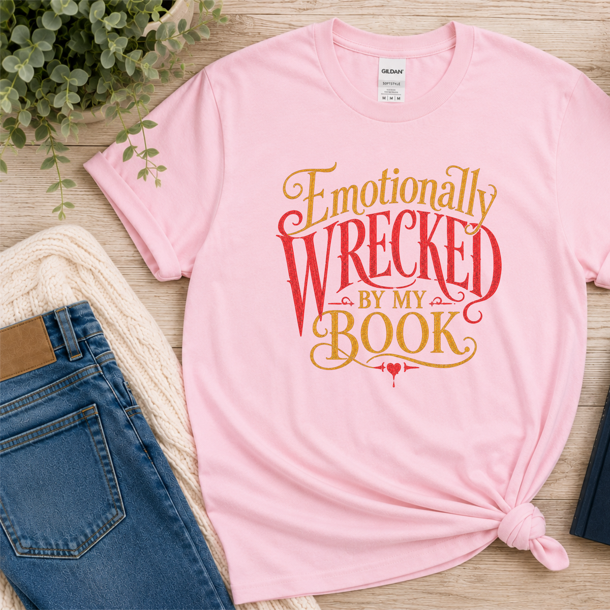Light pink T-shirt laid flat with ornate red and gold lettering and the words “Emotionally Wrecked by My Book.”