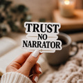 Hand holding a white-bordered sticker reading “Trust No Narrator,” featuring black serif lettering with gold lines and a small star.