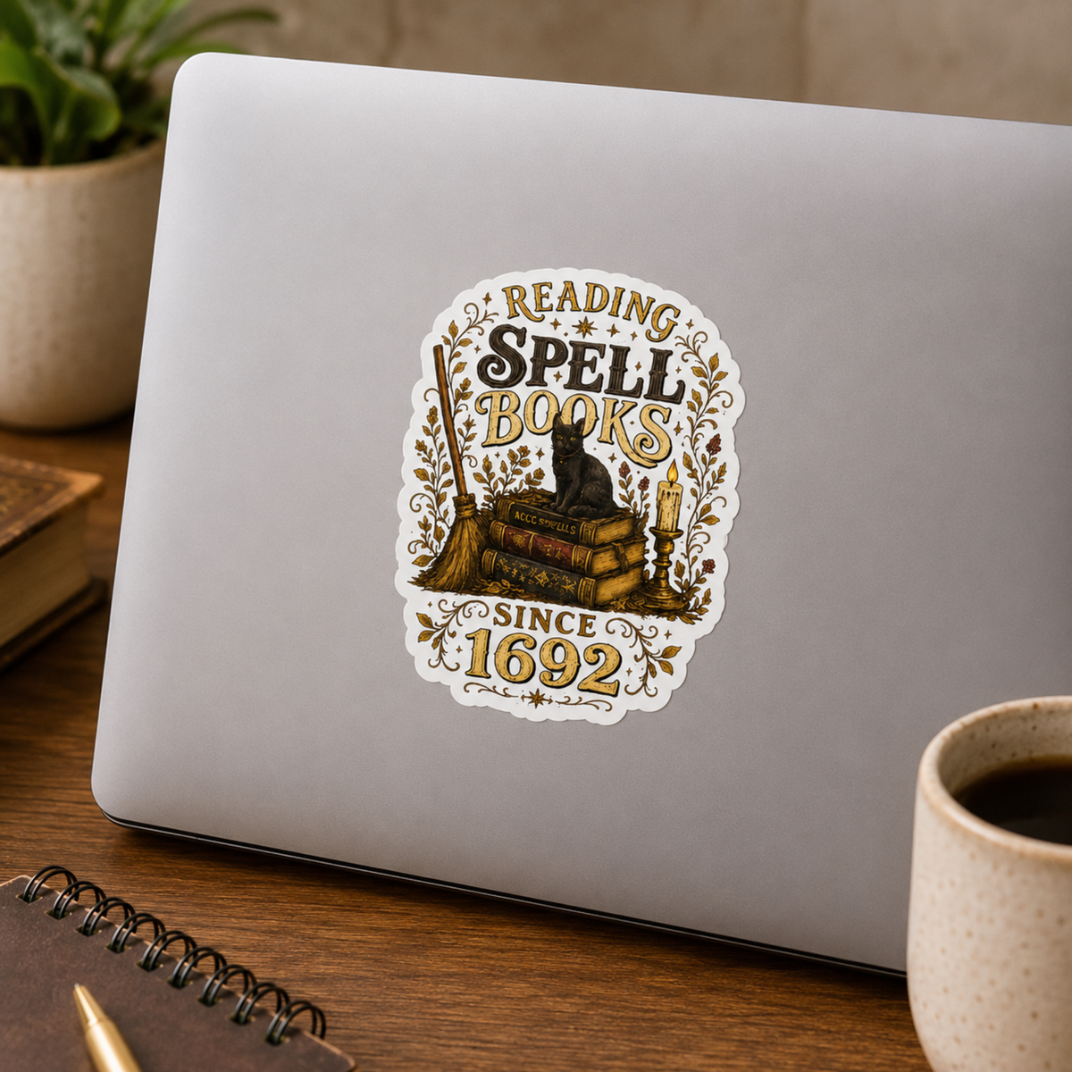 “Reading Spell Books Since 1692” sticker with black and gold lettering, a black cat on antique books, a broom, candle, and botanical flourishes, shown on a silver laptop.