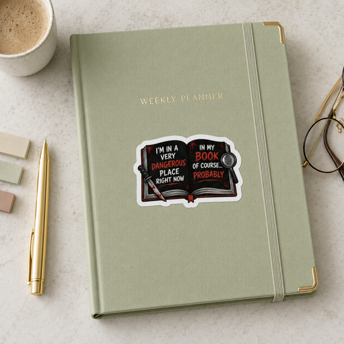 “I’m in a very dangerous place right now. In my book, of course… probably” sticker with an open black book with red and white lettering, a bloodstained knife, and a magnifying glass, shown on a sage weekly planner.