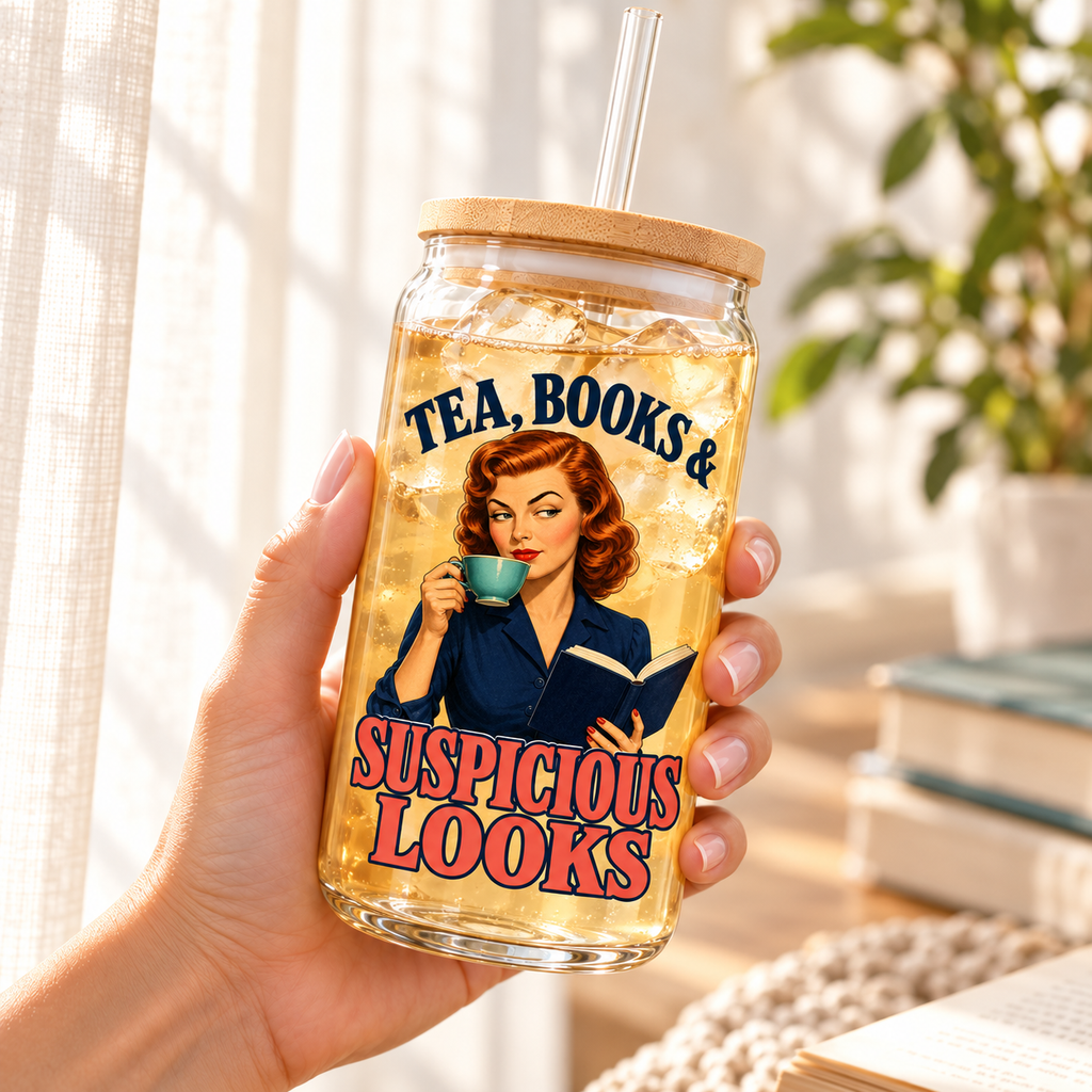 Hand holding an angled “Tea, Books & Suspicious Looks” glass sipper of light iced tea near a curtain; retro red-haired reader artwork, bamboo lid and clear straw.
