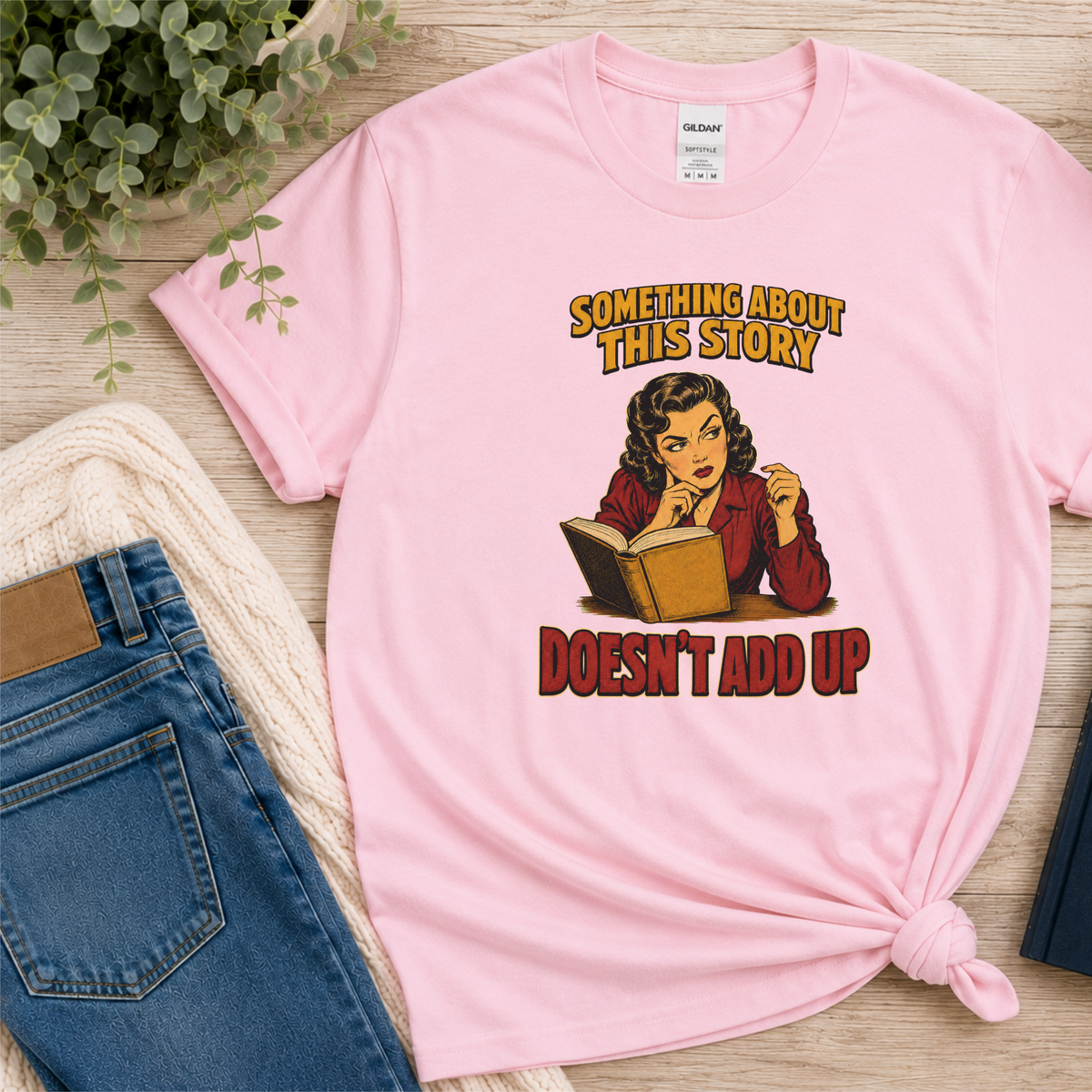 Light pink T-shirt laid flat with an illustrated reader holding an open book and the words “Something About This Story Doesn’t Add Up.”