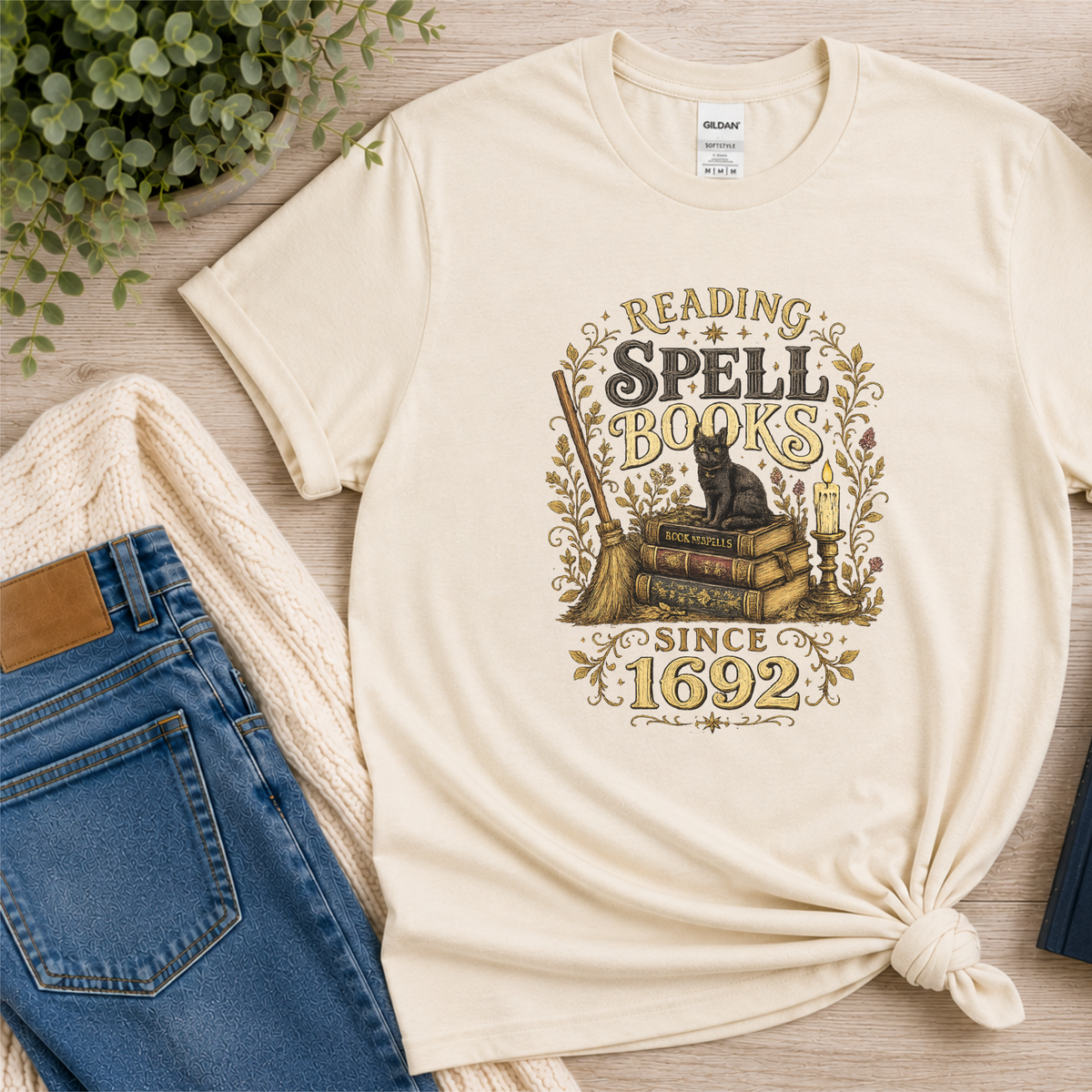Natural cream T-shirt laid flat with a black cat on stacked books with candles and botanical flourishes and the words “Reading Spell Books Since 1692.”