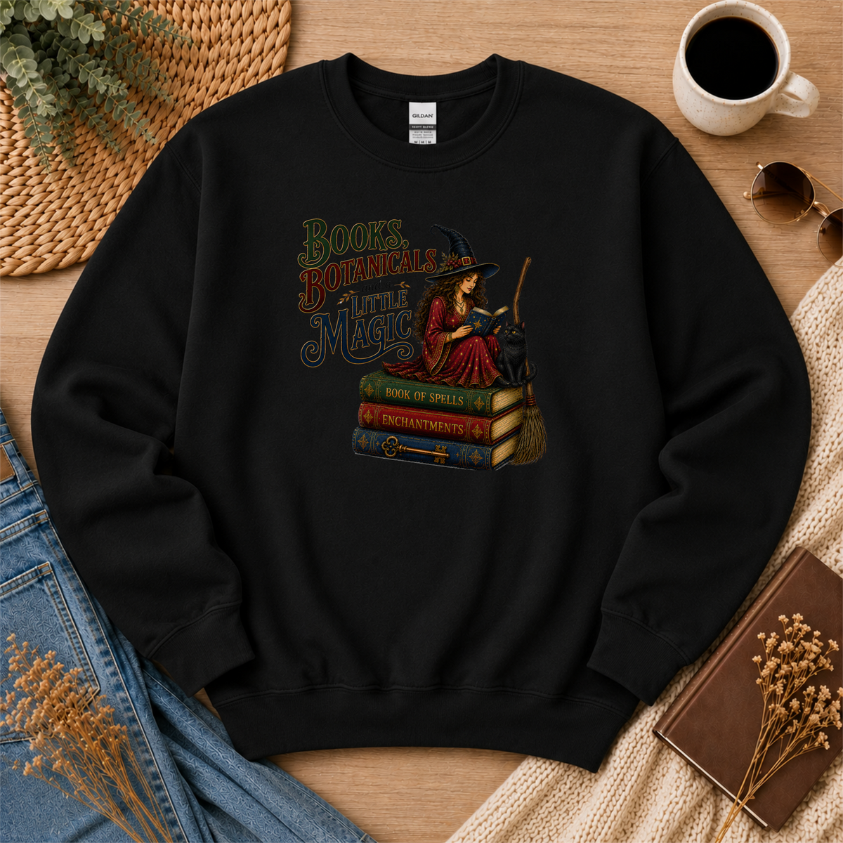 Black crewneck sweatshirt laid flat with a witch reading on stacked spell books beside a black cat and broom and the words “Books, Botanicals and a Little Magic.”