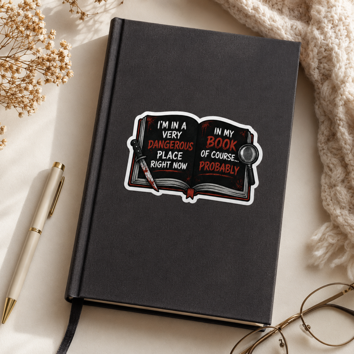 “I’m in a very dangerous place right now. In my book, of course… probably” sticker with an open black book with red and white lettering, a bloodstained knife, and a magnifying glass, shown on a black journal.