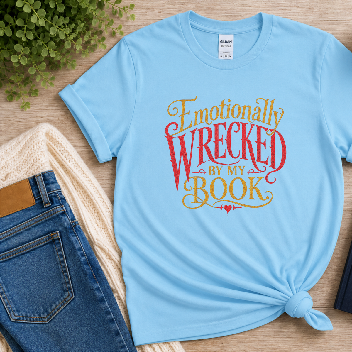 Light blue T-shirt laid flat with ornate red and gold lettering and the words “Emotionally Wrecked by My Book.”