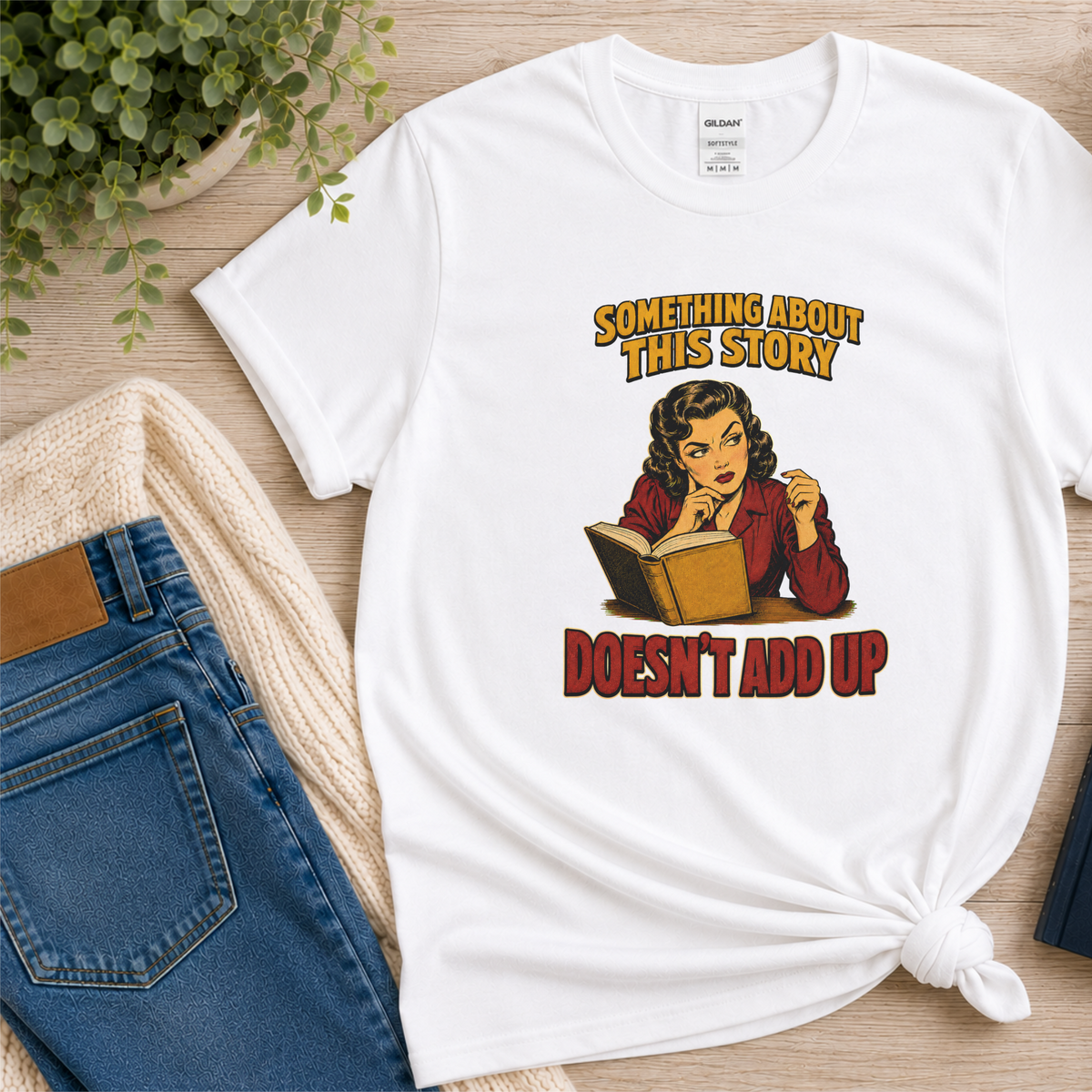 White T-shirt laid flat with an illustrated reader holding an open book and the words “Something About This Story Doesn’t Add Up.”