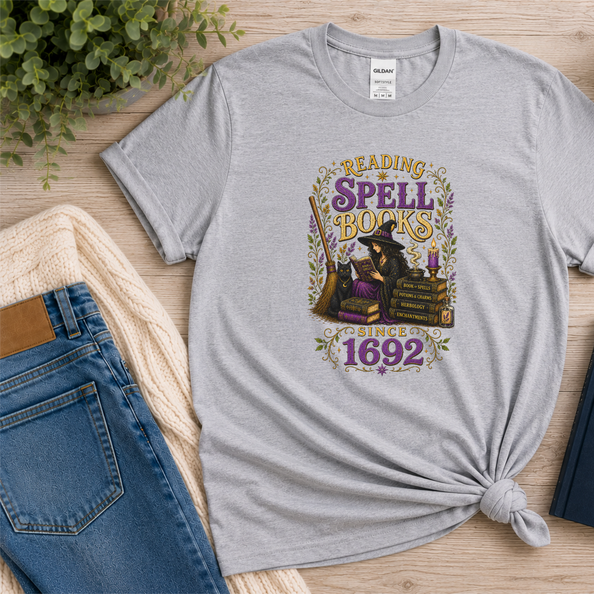 Sport gray T-shirt laid flat with a witch reading beside stacked books and candles and the words “Reading Spell Books Since 1692.”