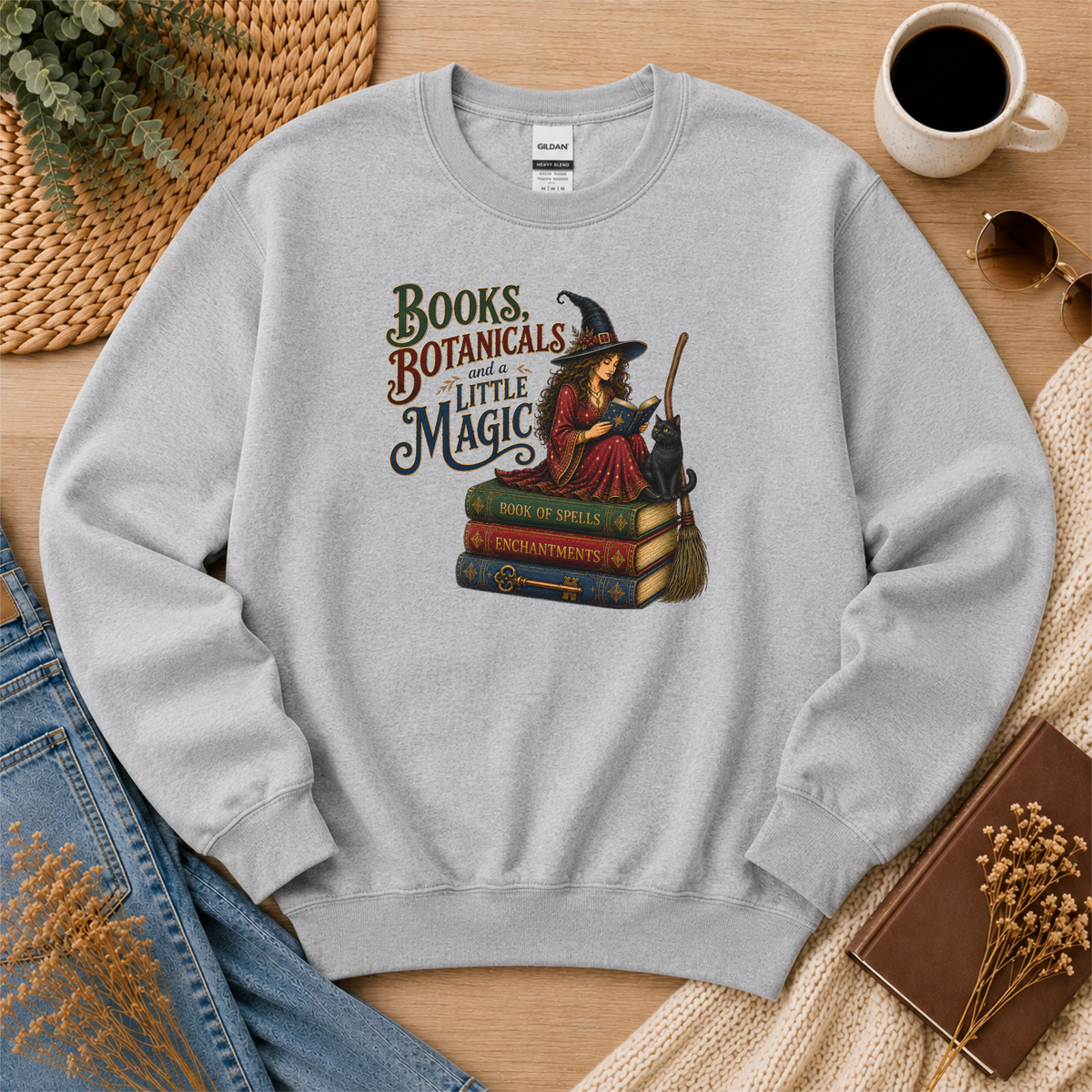 Sport gray crewneck sweatshirt laid flat with a witch reading on stacked spell books beside a black cat and broom and the words “Books, Botanicals and a Little Magic.”