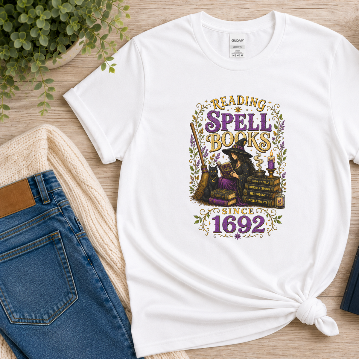 White T-shirt laid flat with a witch reading beside stacked books and candles and the words “Reading Spell Books Since 1692.”