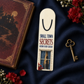 Aluminum page-holder bookmark reading “Small Town Secrets Behind Every Curtain,” with a silhouetted figure behind a glowing blue-curtained window and red flowers, beside a vintage book and key.