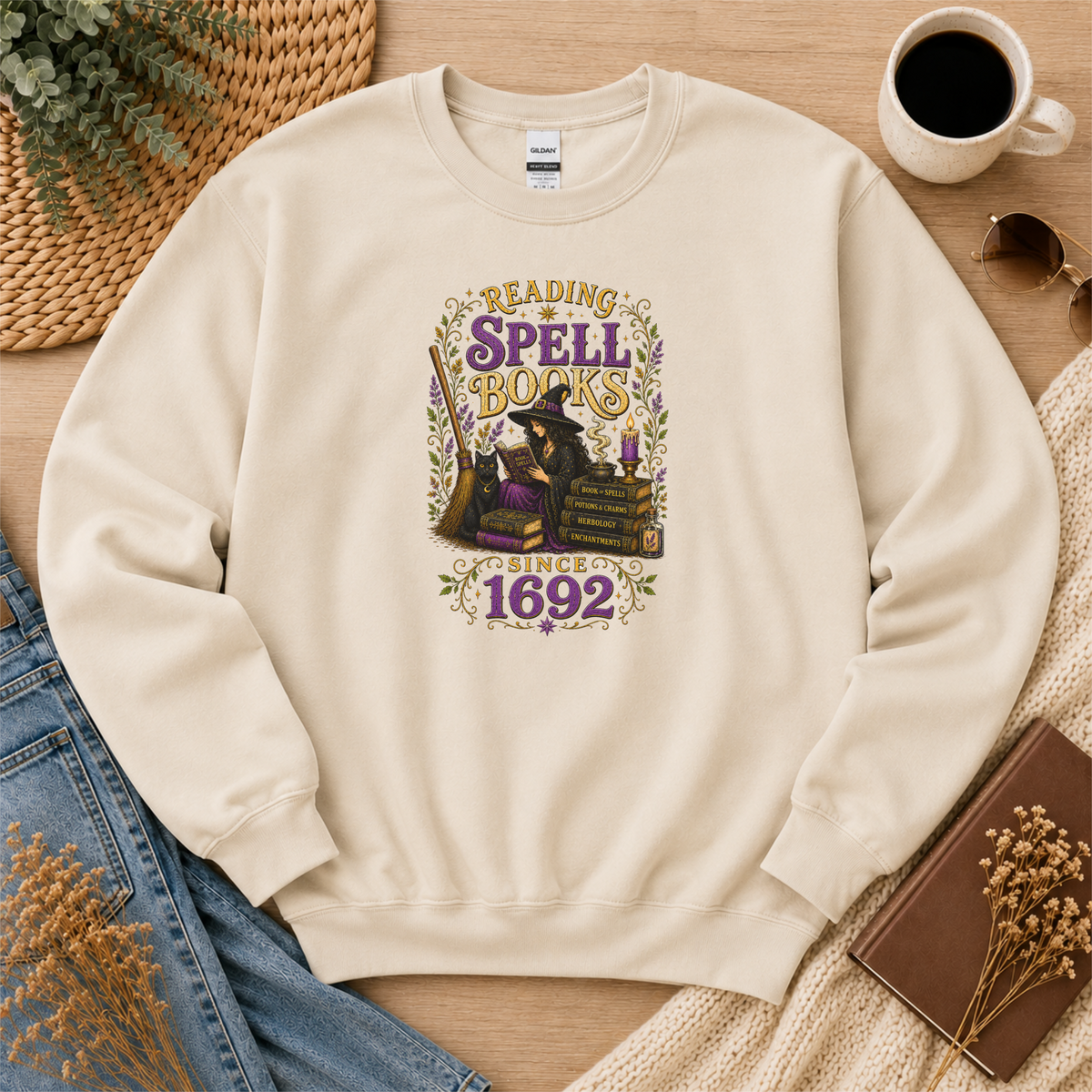 Sand crewneck sweatshirt laid flat with a witch reading beside stacked books and candles and the words “Reading Spell Books Since 1692.”