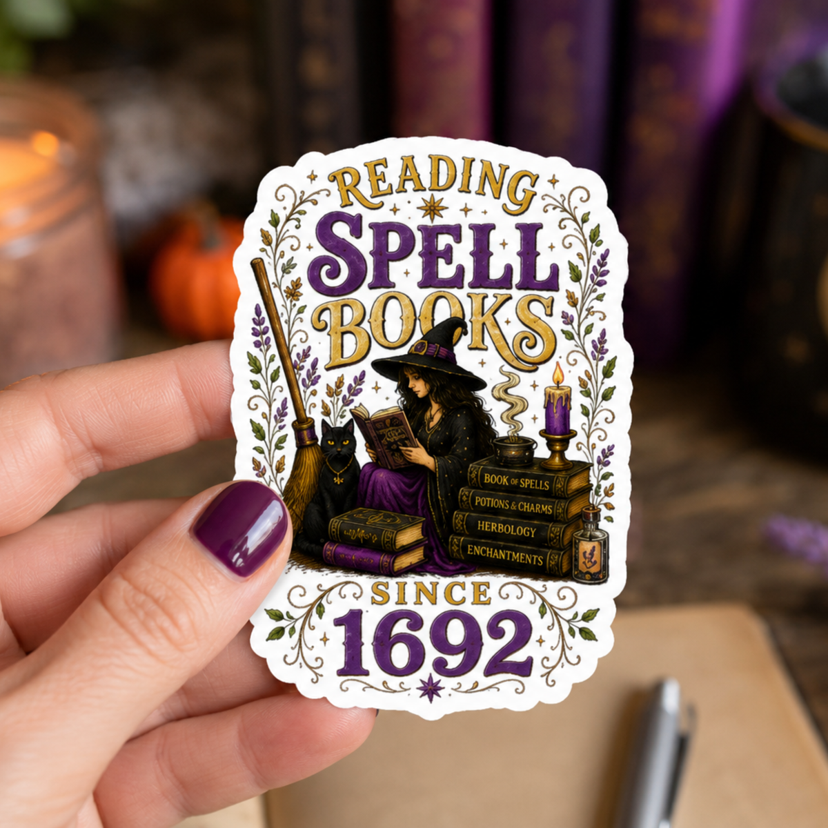 Hand holding a white-bordered sticker reading “Reading Spell Books Since 1692,” featuring purple and gold lettering, a witch reading beside a black cat, candle, broom, and stacked spell books.