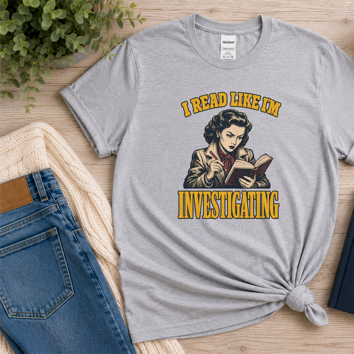 Sport gray T-shirt laid flat with an illustrated reader holding a book and pen and the words “I Read Like I’m Investigating.”