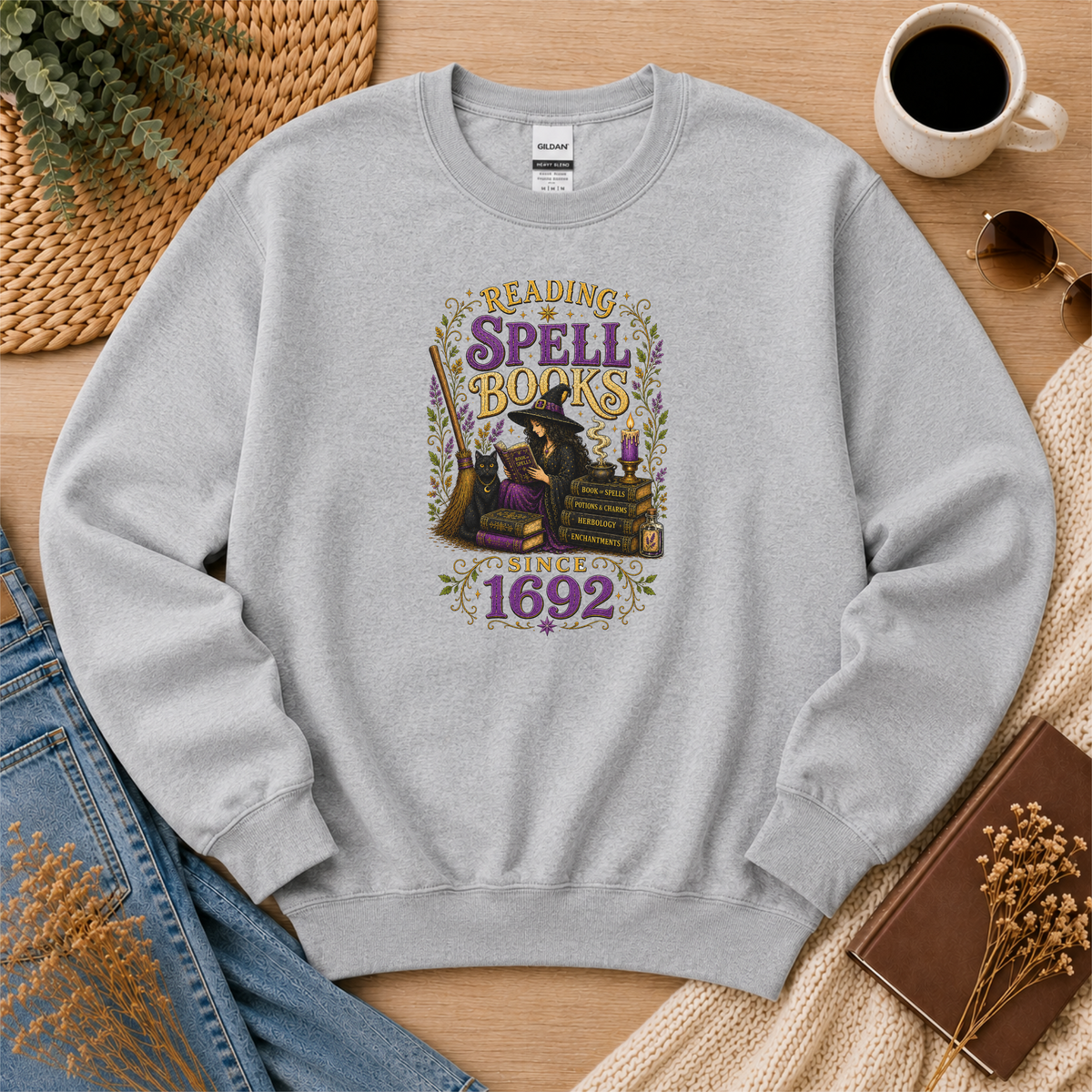 Sport gray crewneck sweatshirt laid flat with a witch reading beside stacked books and candles and the words “Reading Spell Books Since 1692.”