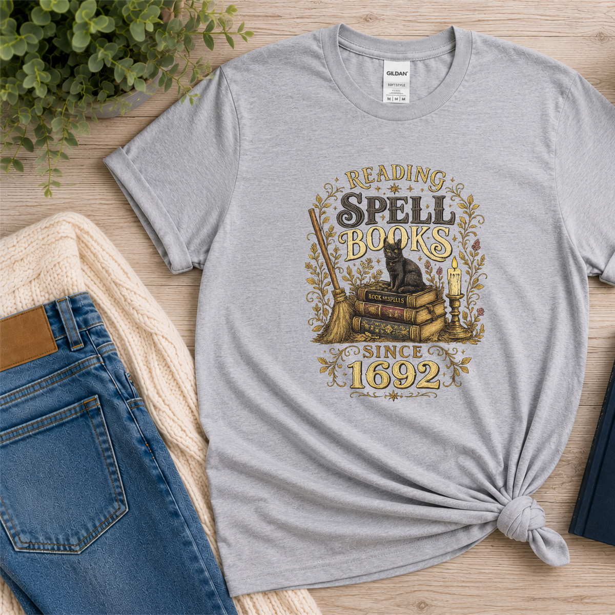 Sport gray T-shirt laid flat with a black cat on stacked books with candles and botanical flourishes and the words “Reading Spell Books Since 1692.”