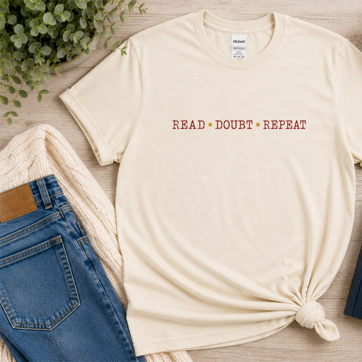 Natural cream T-shirt laid flat with small red lettering across the chest and the words “Read Doubt Repeat.”
