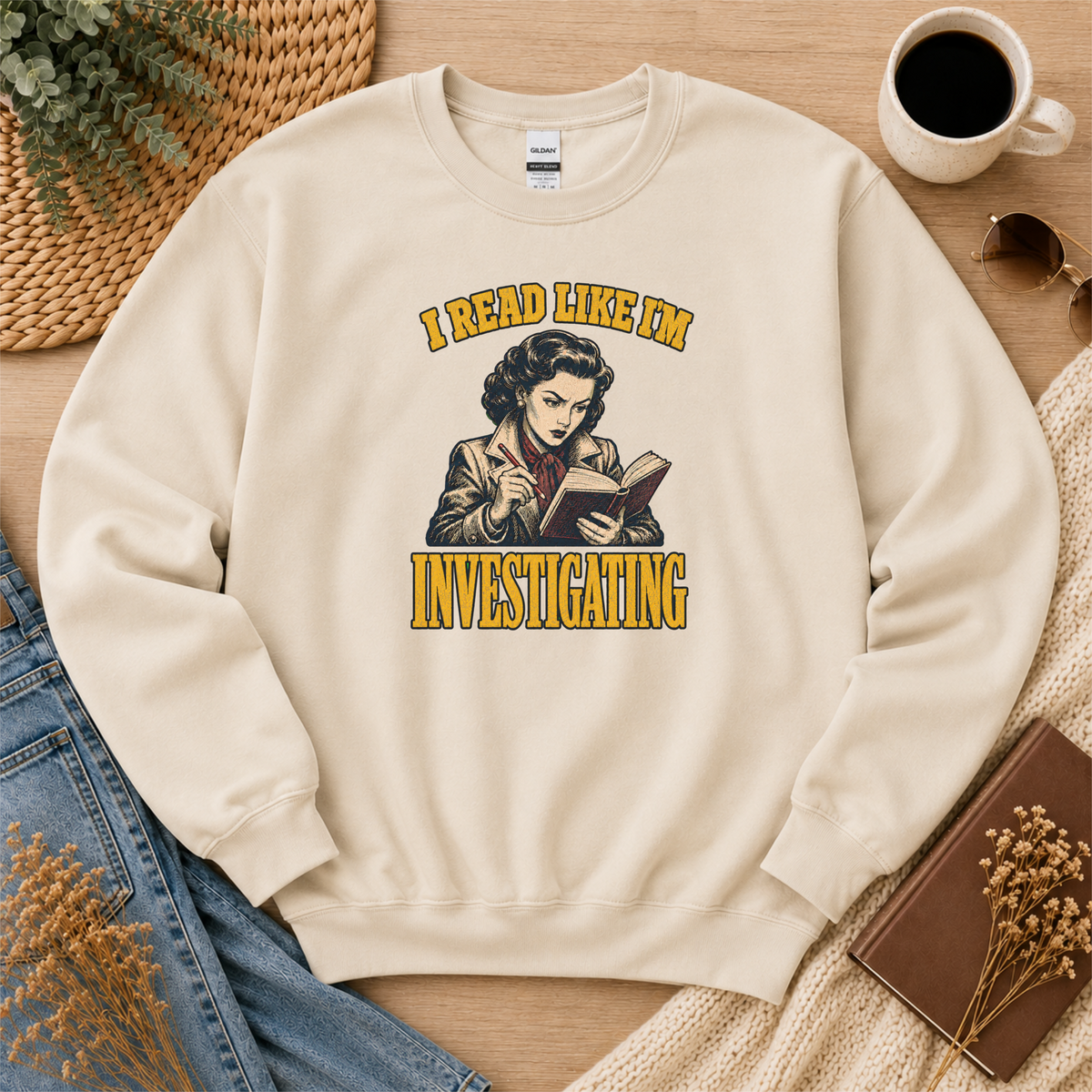 Sand crewneck sweatshirt laid flat with an illustrated reader holding a book and pen and the words “I Read Like I’m Investigating.”