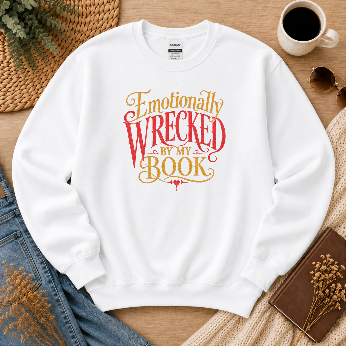 White crewneck sweatshirt laid flat with ornate red and gold lettering and the words “Emotionally Wrecked by My Book.”
