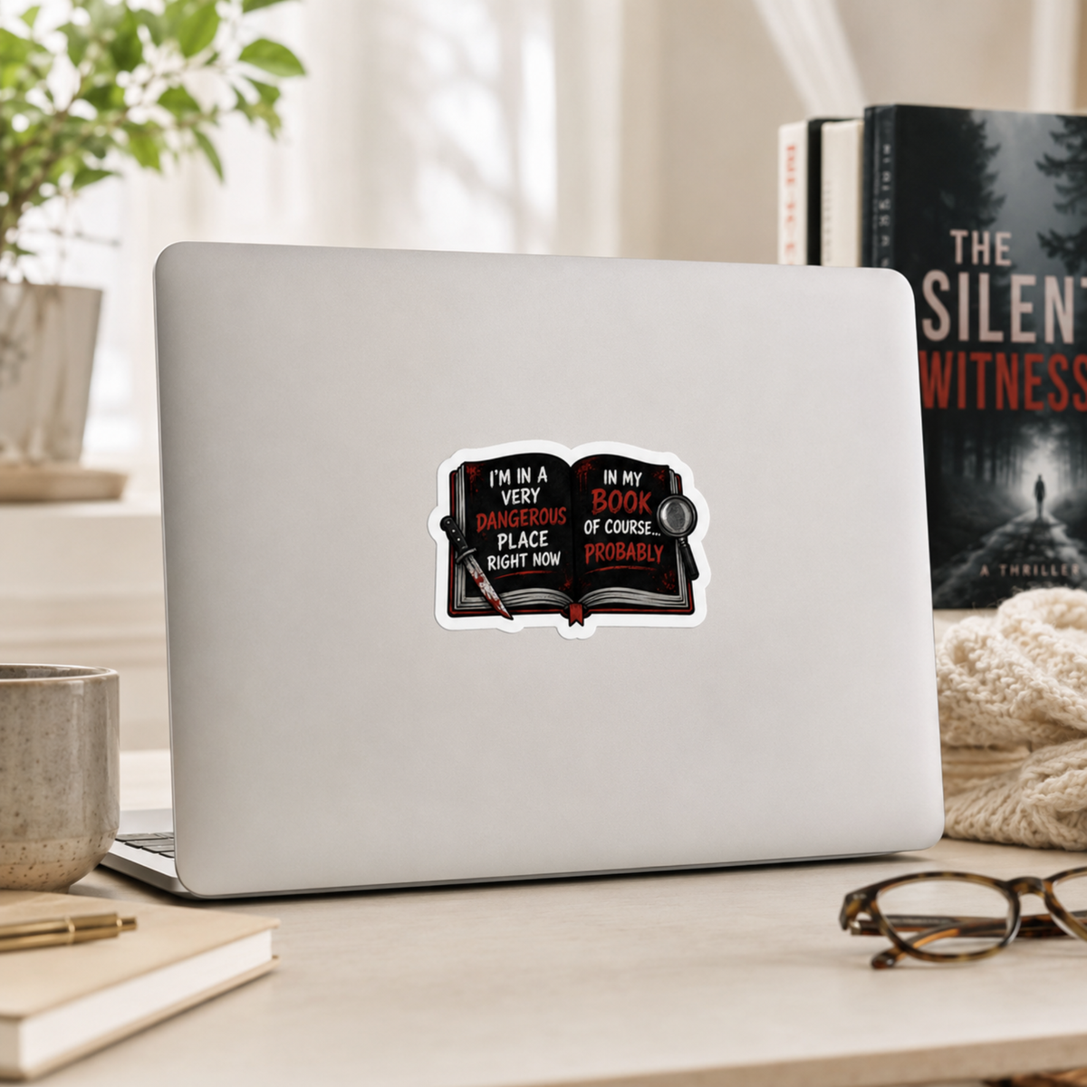 “I’m in a very dangerous place right now. In my book, of course… probably” sticker with an open black book with red and white lettering, a bloodstained knife, and a magnifying glass, shown on a silver laptop.