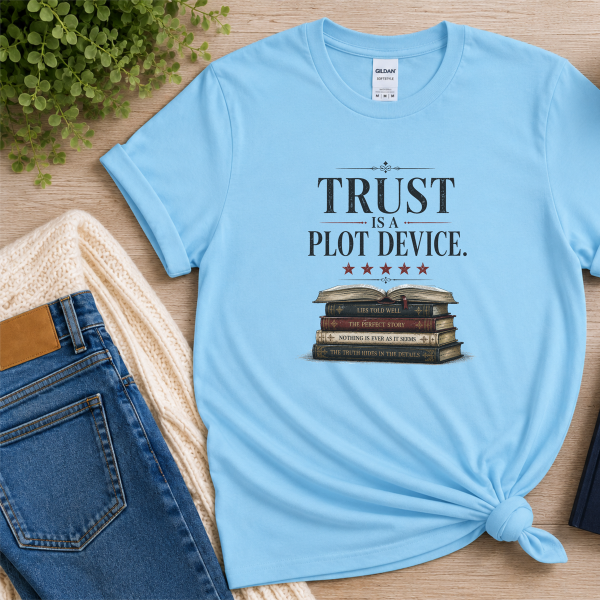 Light blue T-shirt laid flat with stars above a stack of books and the words “Trust Is a Plot Device.”