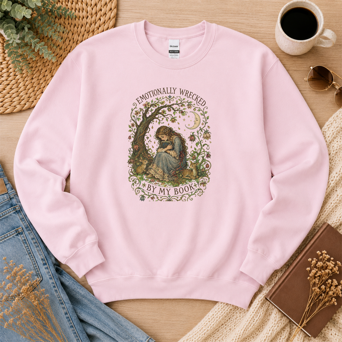 Light pink crewneck sweatshirt laid flat with a reader beneath a tree, flowers and a crescent moon and the words “Emotionally Wrecked by My Book.”
