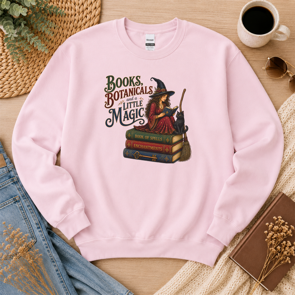 Light pink crewneck sweatshirt laid flat with a witch reading on stacked spell books beside a black cat and broom and the words “Books, Botanicals and a Little Magic.”