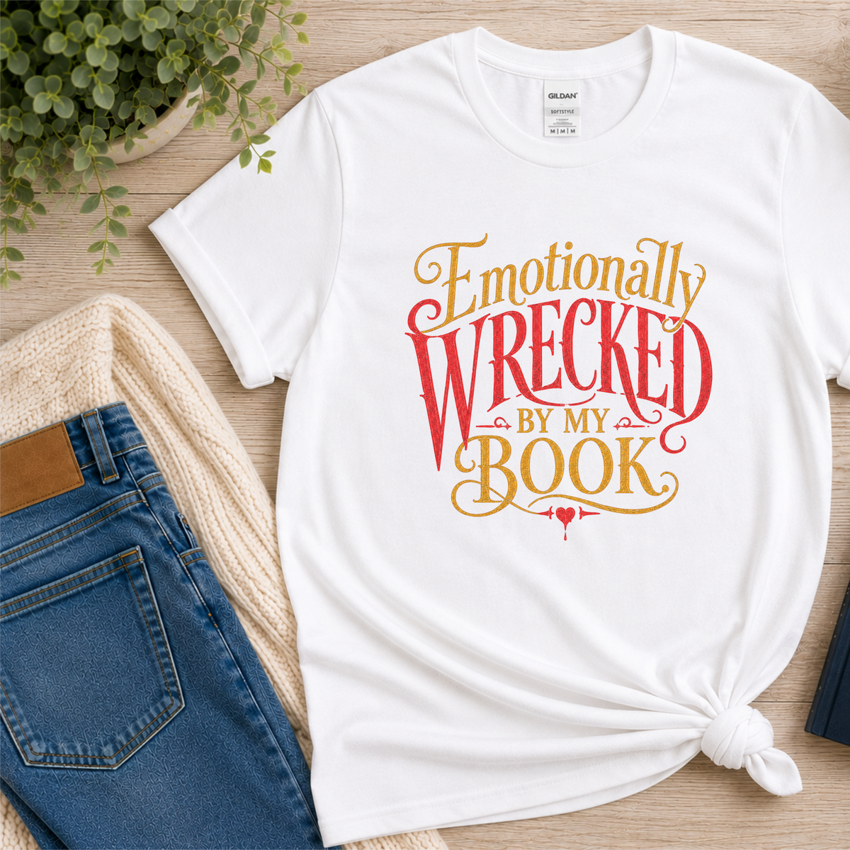White T-shirt laid flat with ornate red and gold lettering and the words “Emotionally Wrecked by My Book.”