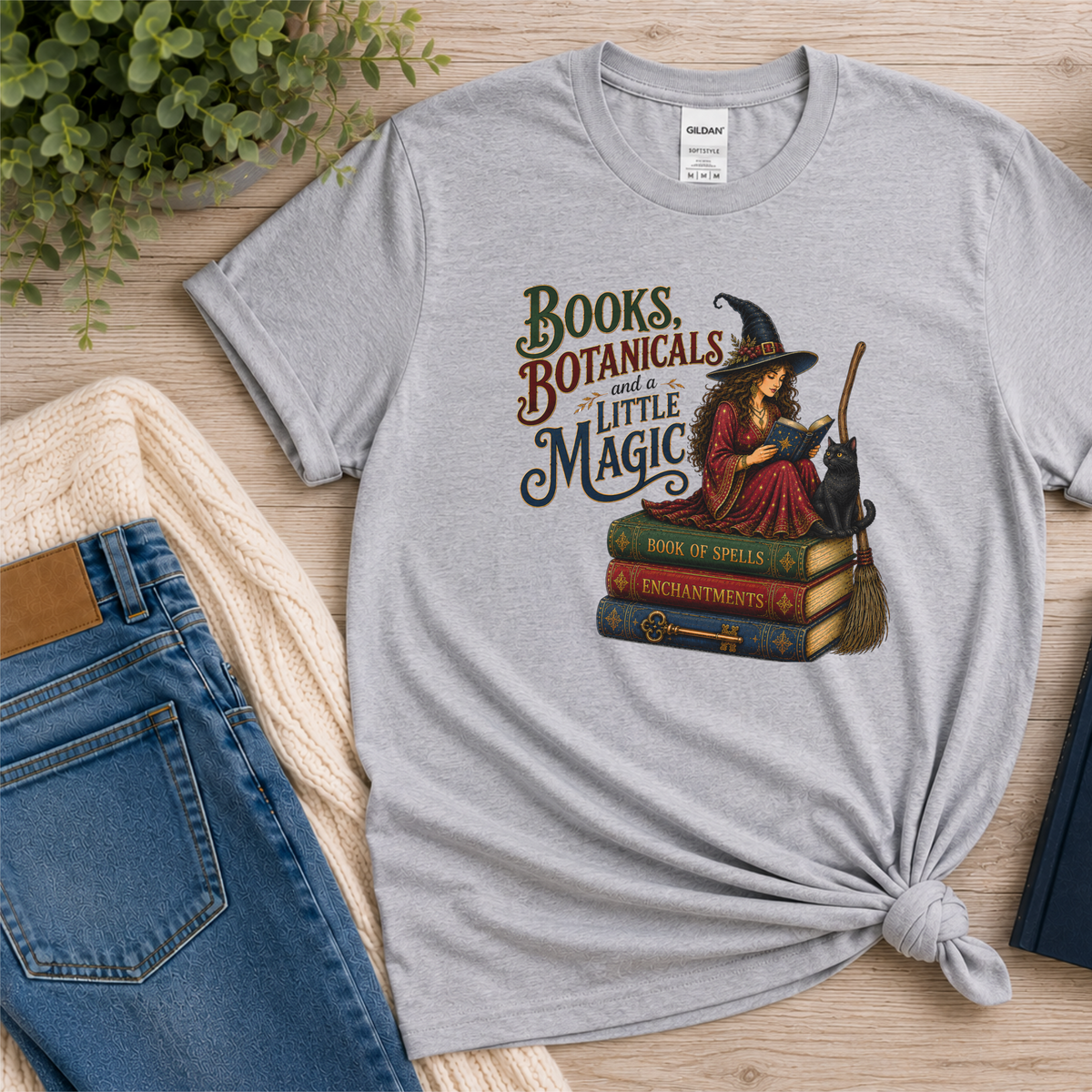 Sport gray T-shirt laid flat with a witch reading on stacked spell books beside a black cat and broom and the words “Books, Botanicals and a Little Magic.”