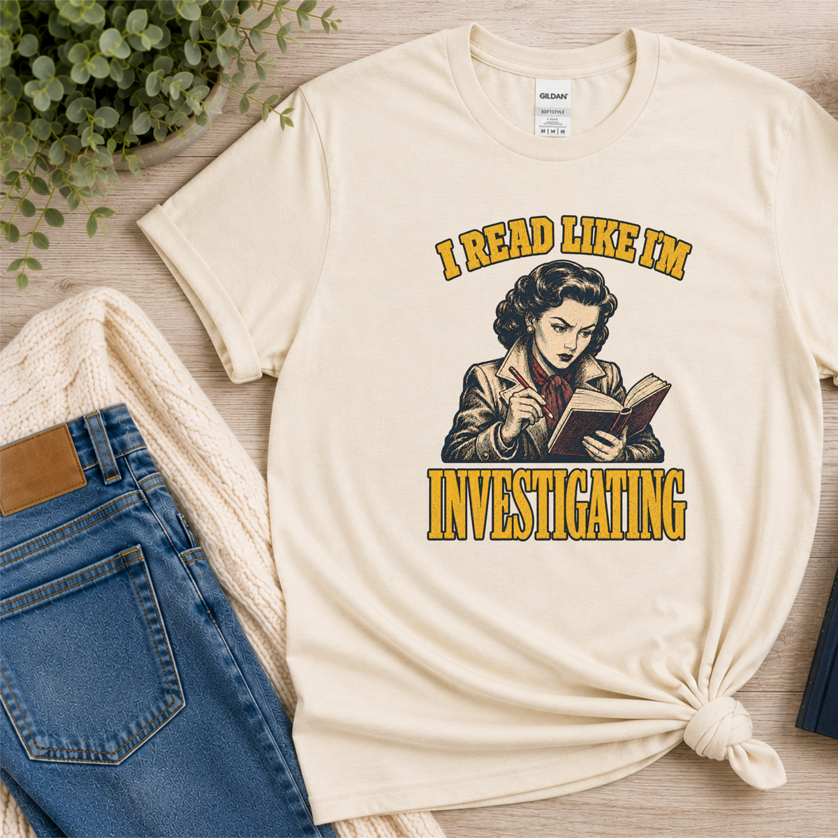 Natural cream T-shirt laid flat with an illustrated reader holding a book and pen and the words “I Read Like I’m Investigating.”