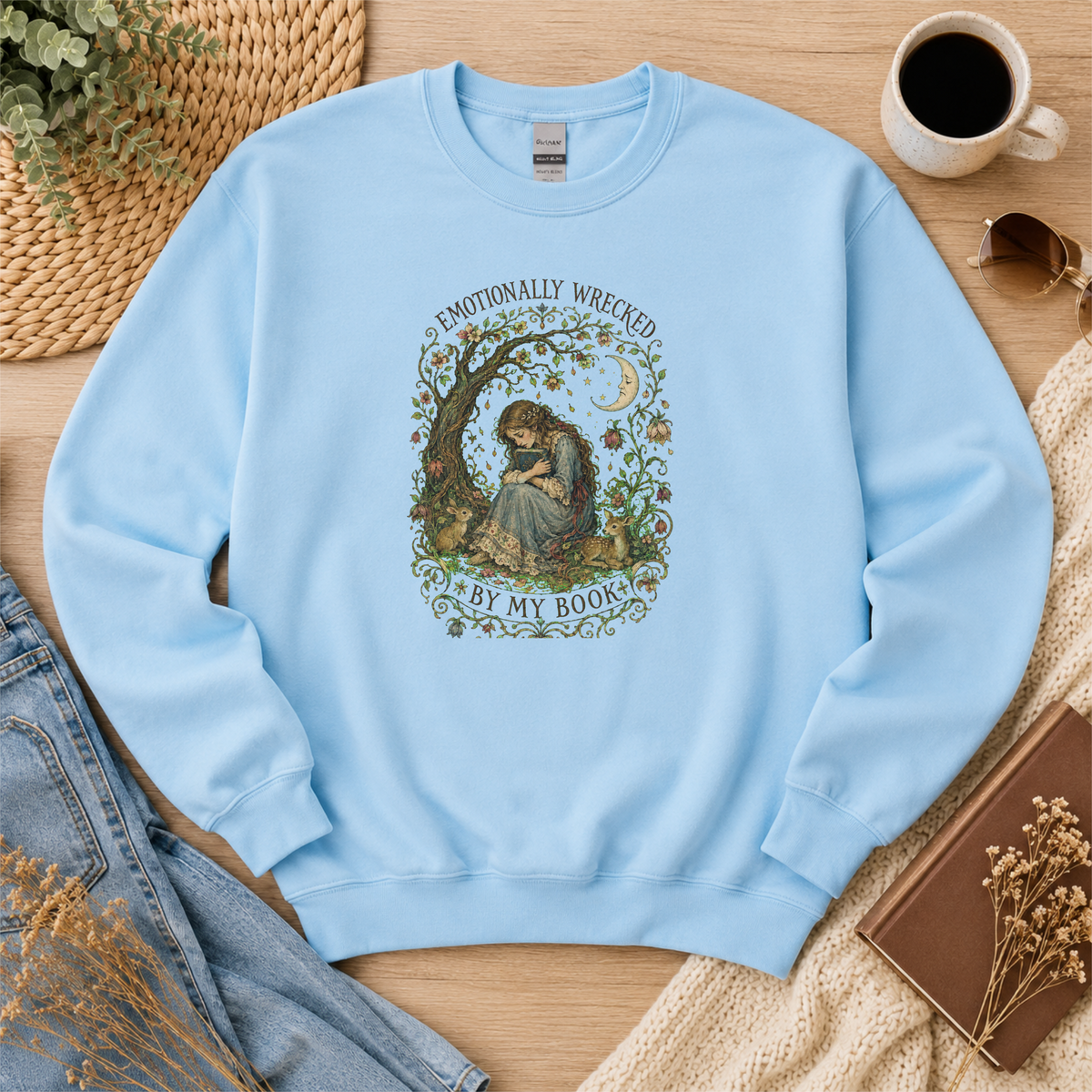Light blue crewneck sweatshirt laid flat with a reader beneath a tree, flowers and a crescent moon and the words “Emotionally Wrecked by My Book.”
