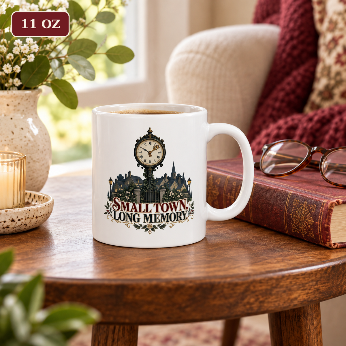 11 oz white mug with the “Small Town, Long Memory” clock-and-town artwork on a wooden side table beside vintage books and reading glasses.