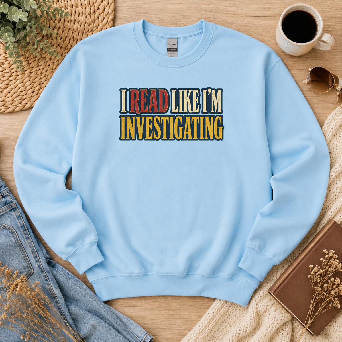 Light blue crewneck sweatshirt laid flat with bold vintage lettering and the words “I Read Like I’m Investigating.”