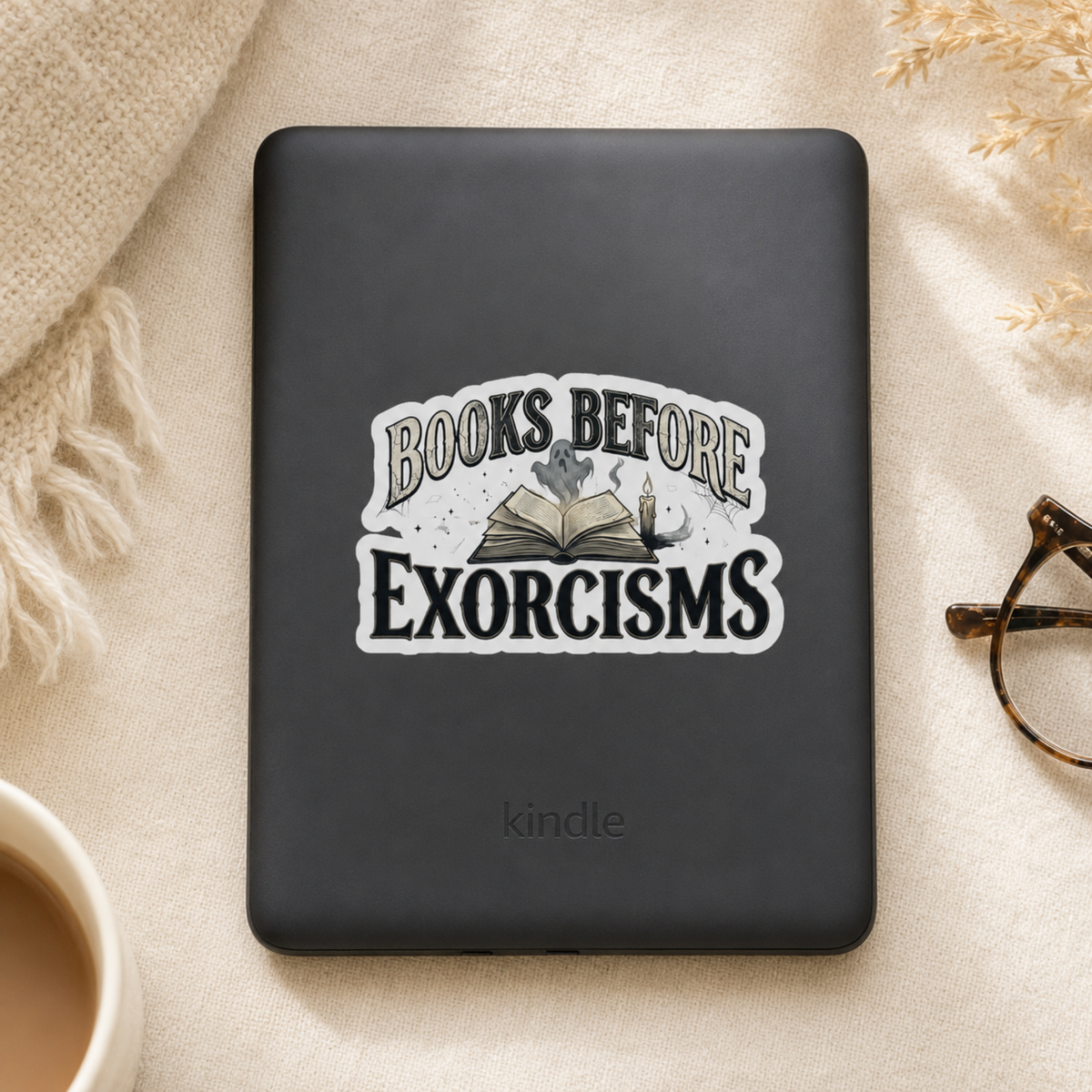 “Books Before Exorcisms” sticker with black lettering, an open book, gray ghost, candle, and cobweb, shown on a black Kindle.