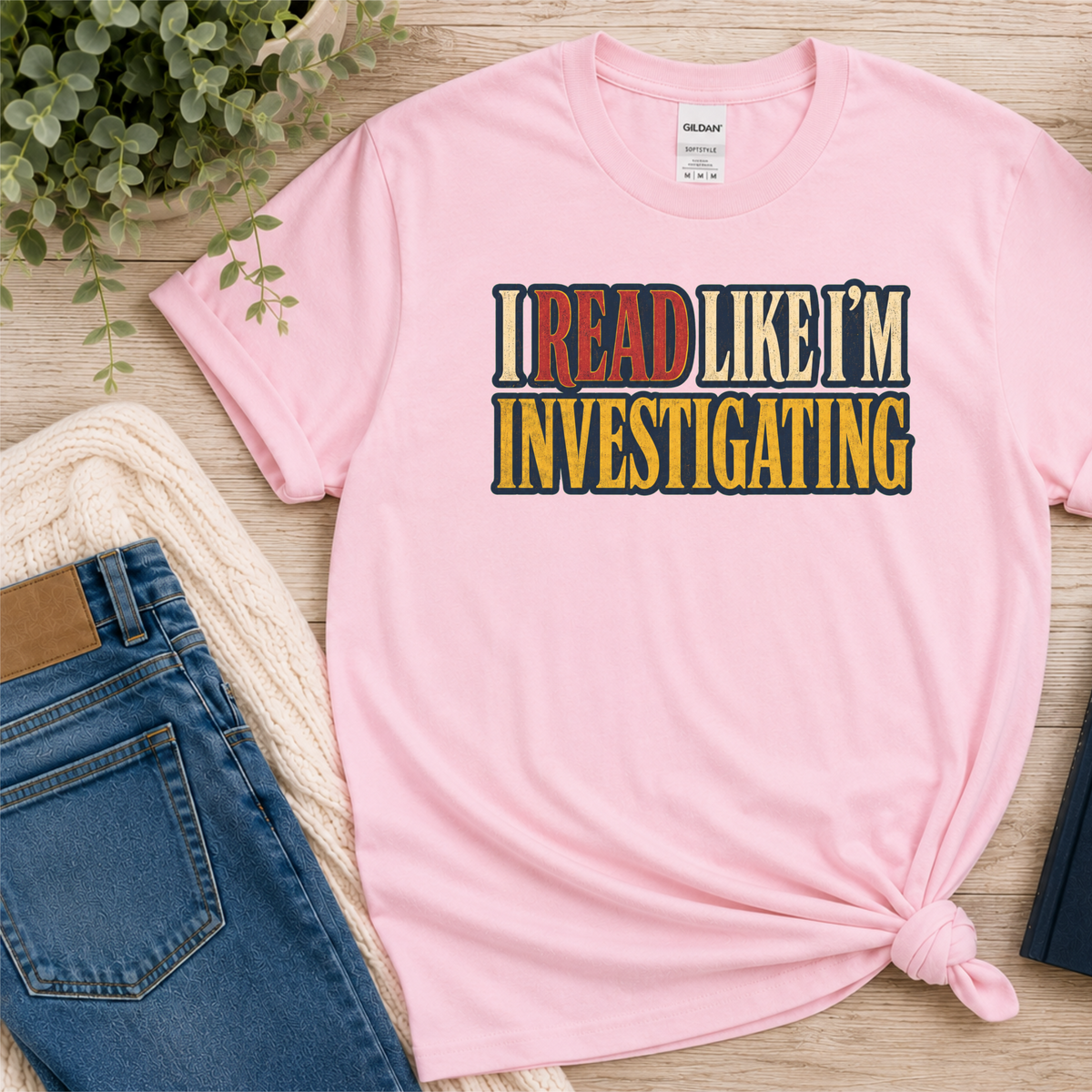 Light pink T-shirt laid flat with bold vintage lettering and the words “I Read Like I’m Investigating.”