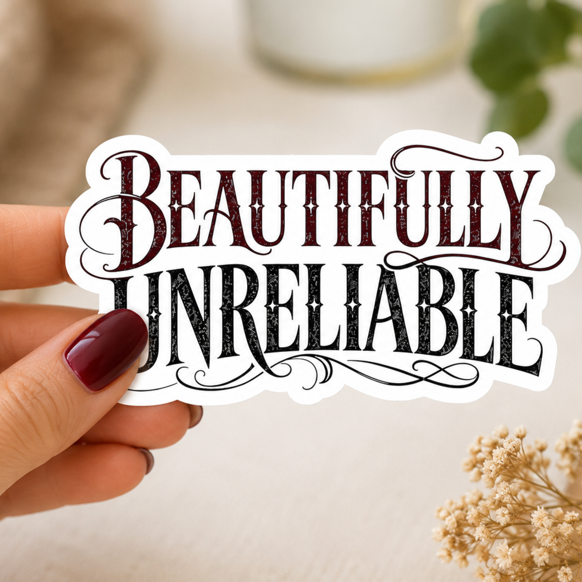 Hand holding a white-bordered sticker reading “Beautifully Unreliable,” featuring burgundy and black ornate typography.