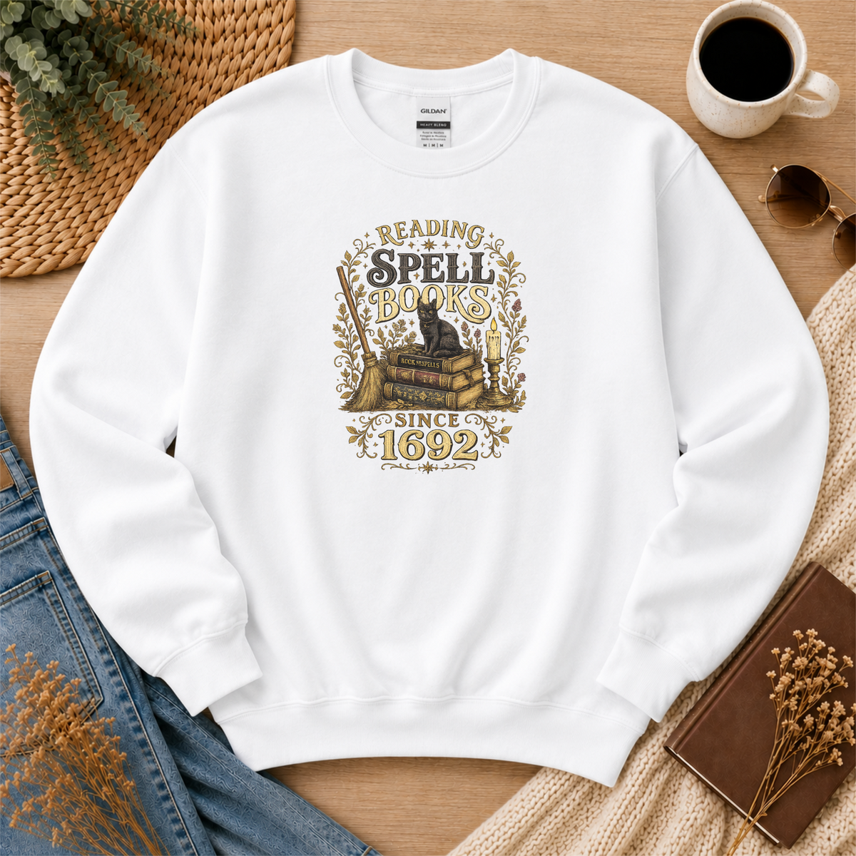 White crewneck sweatshirt laid flat with a black cat on stacked books with candles and botanical flourishes and the words “Reading Spell Books Since 1692.”