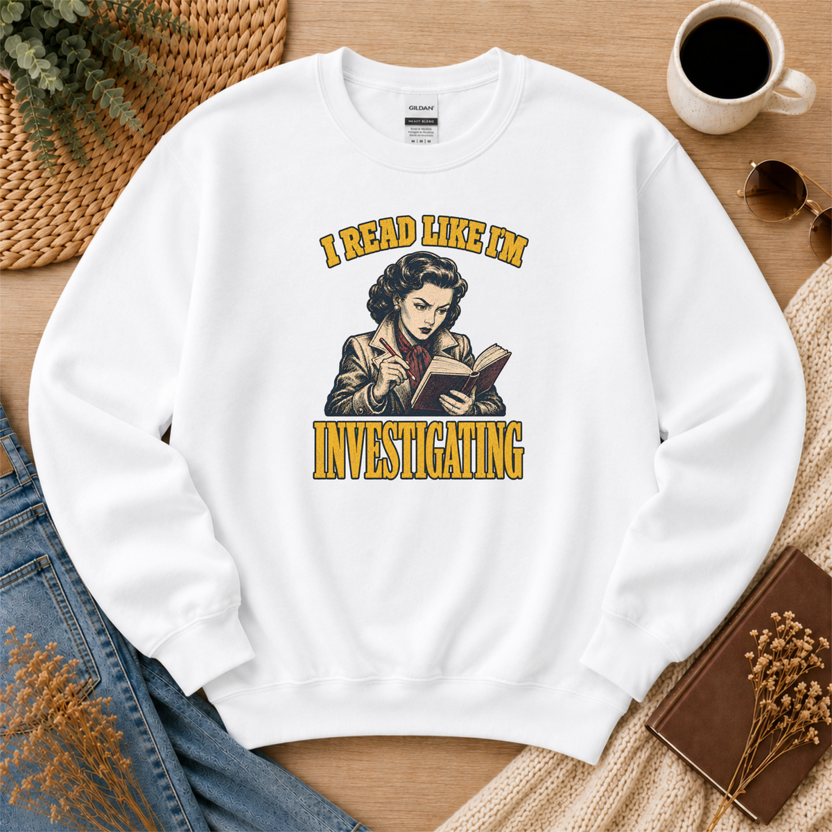 White crewneck sweatshirt laid flat with an illustrated reader holding a book and pen and the words “I Read Like I’m Investigating.”