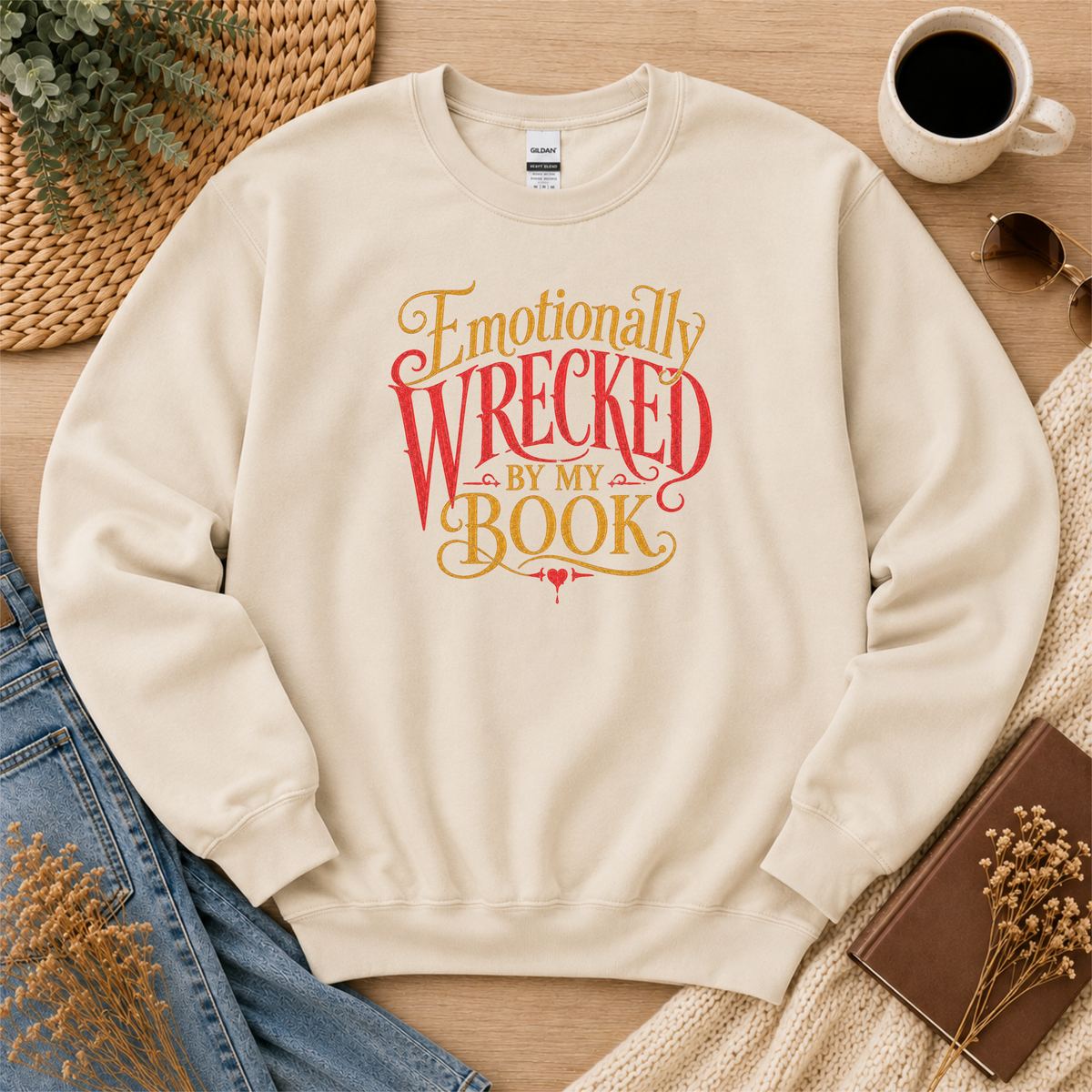 Sand crewneck sweatshirt laid flat with ornate red and gold lettering and the words “Emotionally Wrecked by My Book.”