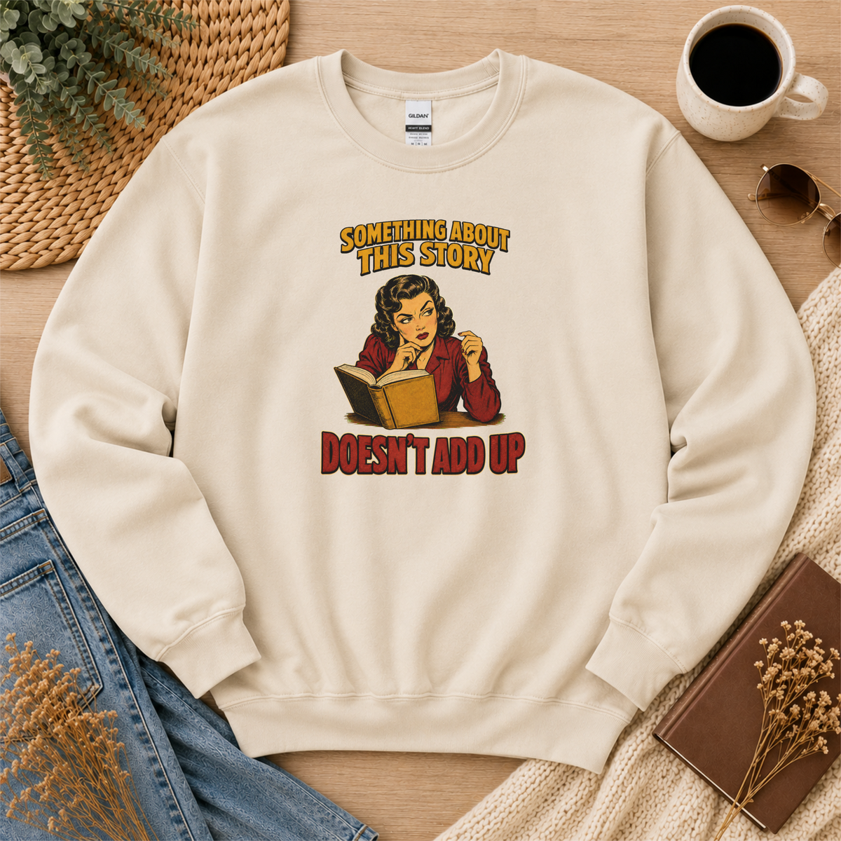 Sand crewneck sweatshirt laid flat with an illustrated reader holding an open book and the words “Something About This Story Doesn’t Add Up.”