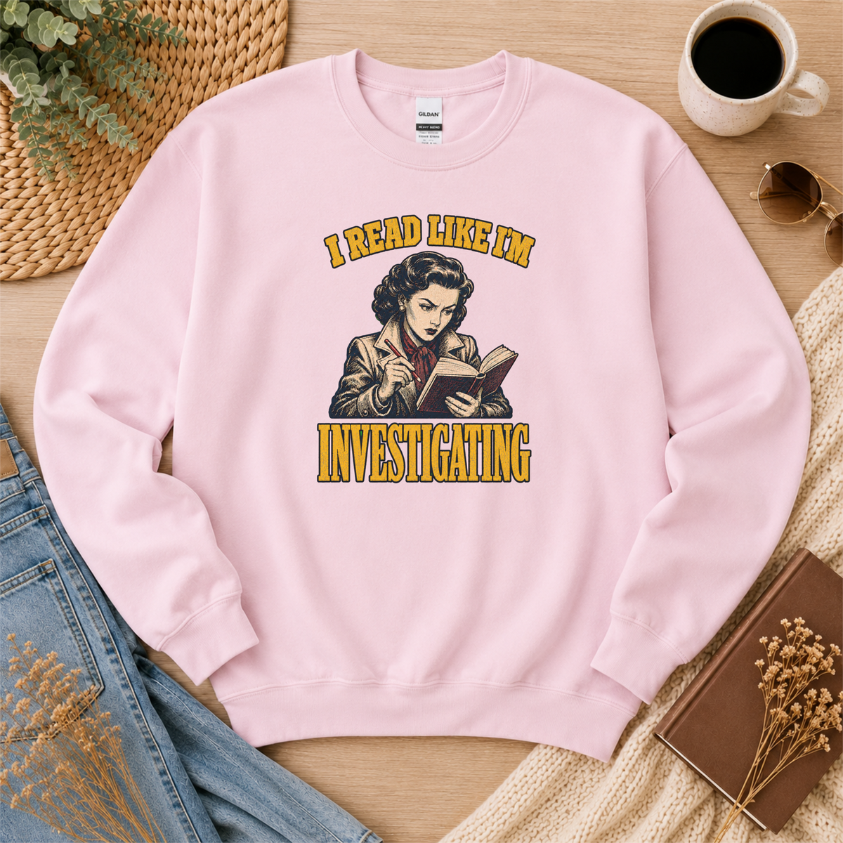 Light pink crewneck sweatshirt laid flat with an illustrated reader holding a book and pen and the words “I Read Like I’m Investigating.”