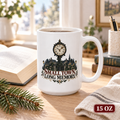 15 oz white ceramic mug featuring a clock above a shadowy town and the words “Small Town, Long Memory,” beside an open book on a sunlit desk.