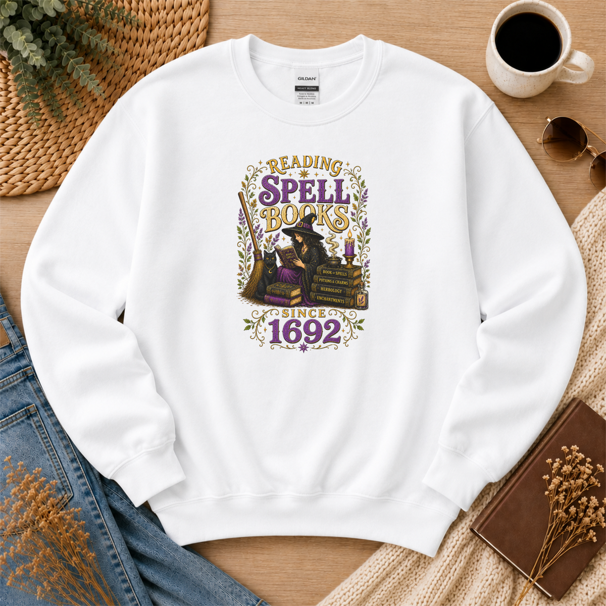 White crewneck sweatshirt laid flat with a witch reading beside stacked books and candles and the words “Reading Spell Books Since 1692.”
