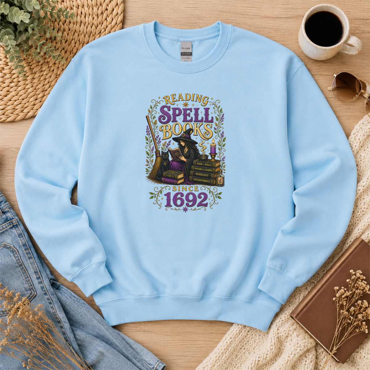 Light blue crewneck sweatshirt laid flat with a witch reading beside stacked books and candles and the words “Reading Spell Books Since 1692.”