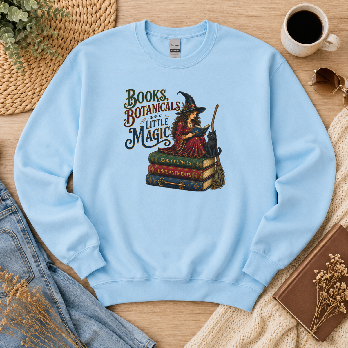 Light blue crewneck sweatshirt laid flat with a witch reading on stacked spell books beside a black cat and broom and the words “Books, Botanicals and a Little Magic.”