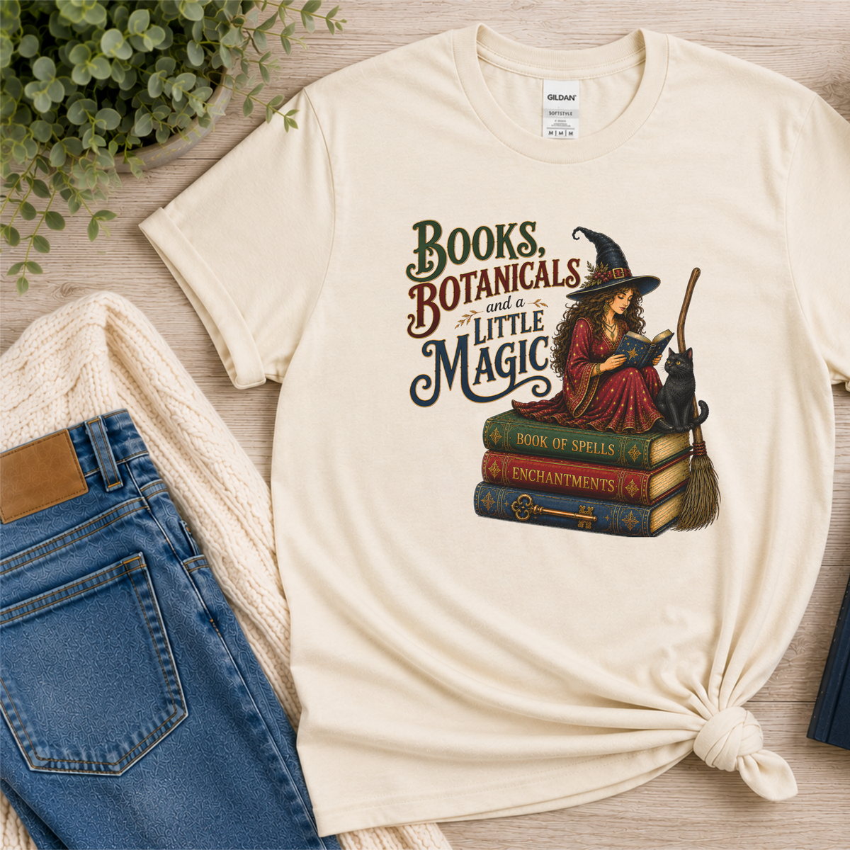 Natural cream T-shirt laid flat with a witch reading on stacked spell books beside a black cat and broom and the words “Books, Botanicals and a Little Magic.”