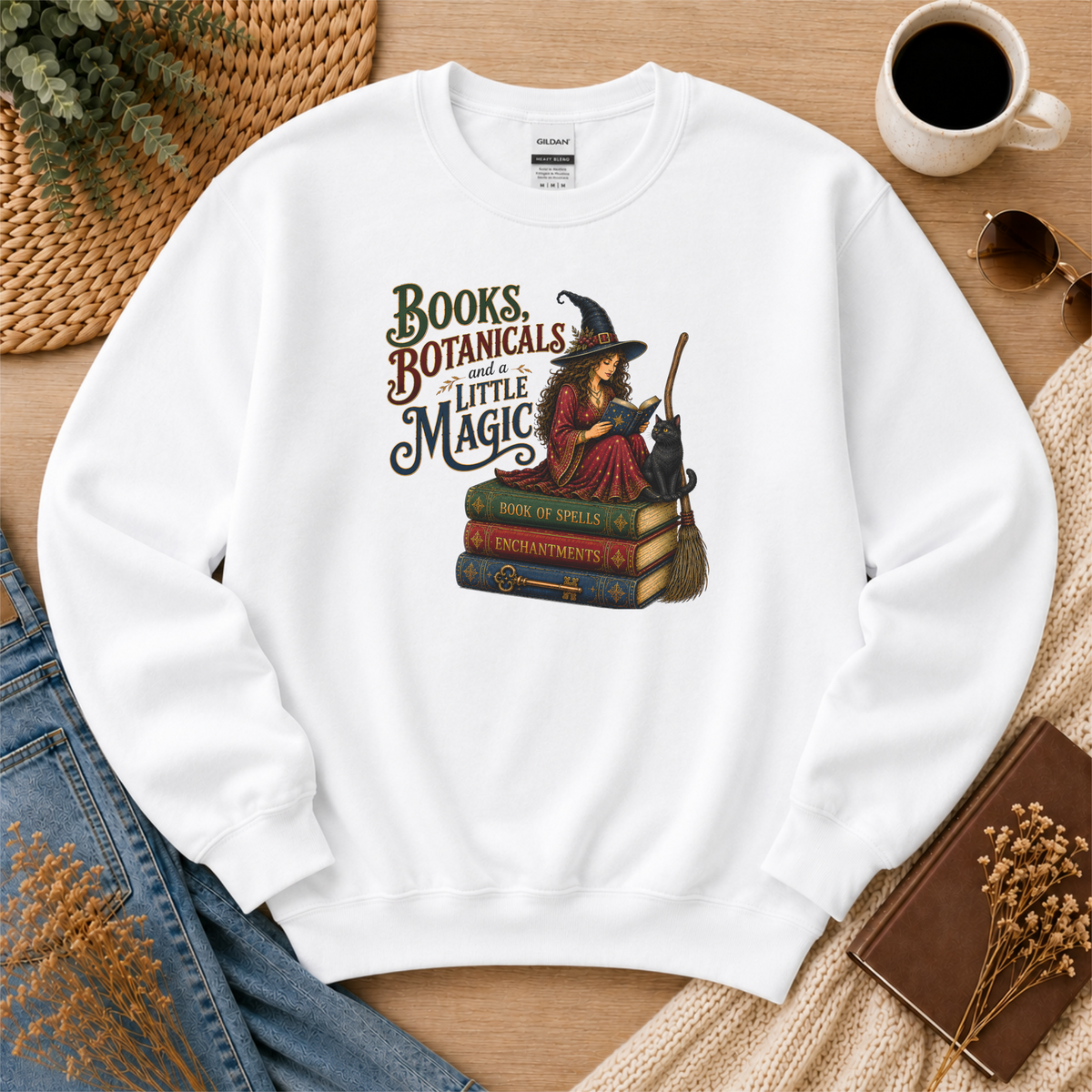 White crewneck sweatshirt laid flat with a witch reading on stacked spell books beside a black cat and broom and the words “Books, Botanicals and a Little Magic.”