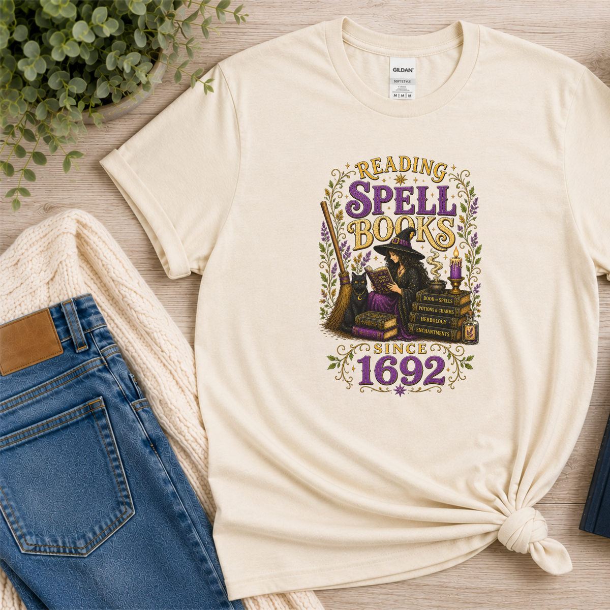 Natural cream T-shirt laid flat with a witch reading beside stacked books and candles and the words “Reading Spell Books Since 1692.”