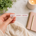 Hand holding a white-bordered sticker reading “Read • Doubt • Repeat,” featuring burgundy typewriter lettering separated by gold dots.